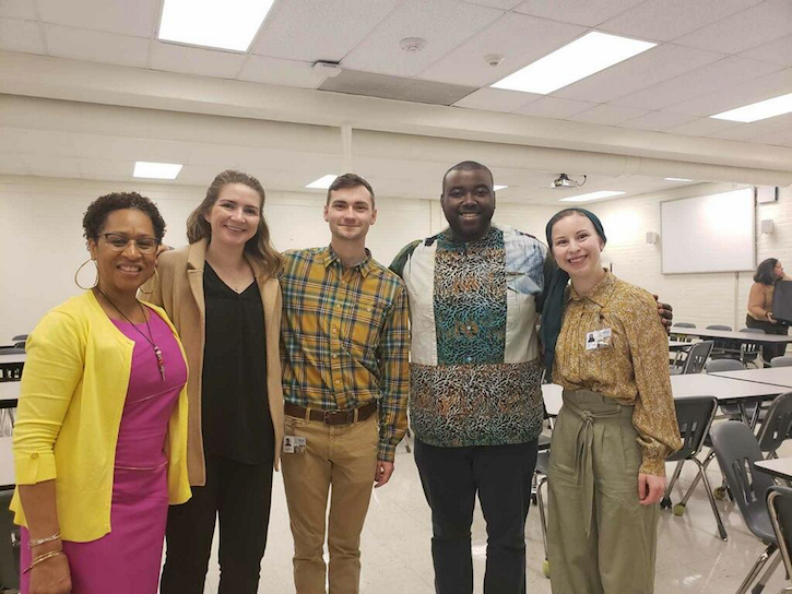 ID: An African American woman, a white woman, a white man, an African American man, and a white women stand side by side smiling at a camera inside a school cafeteria.